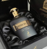 This image shows a perfume bottle of "Amberley Pur Oud" by Maison Alhambra placed inside an open, luxurious box. The bottle is dark, possibly black, with a gold cap and a gold label embossed with the brand name "Amberley." Below the main label, the text "Pur Oud" is written in smaller golden script. The box is lined with black satin fabric, adding an elegant touch to the presentation.