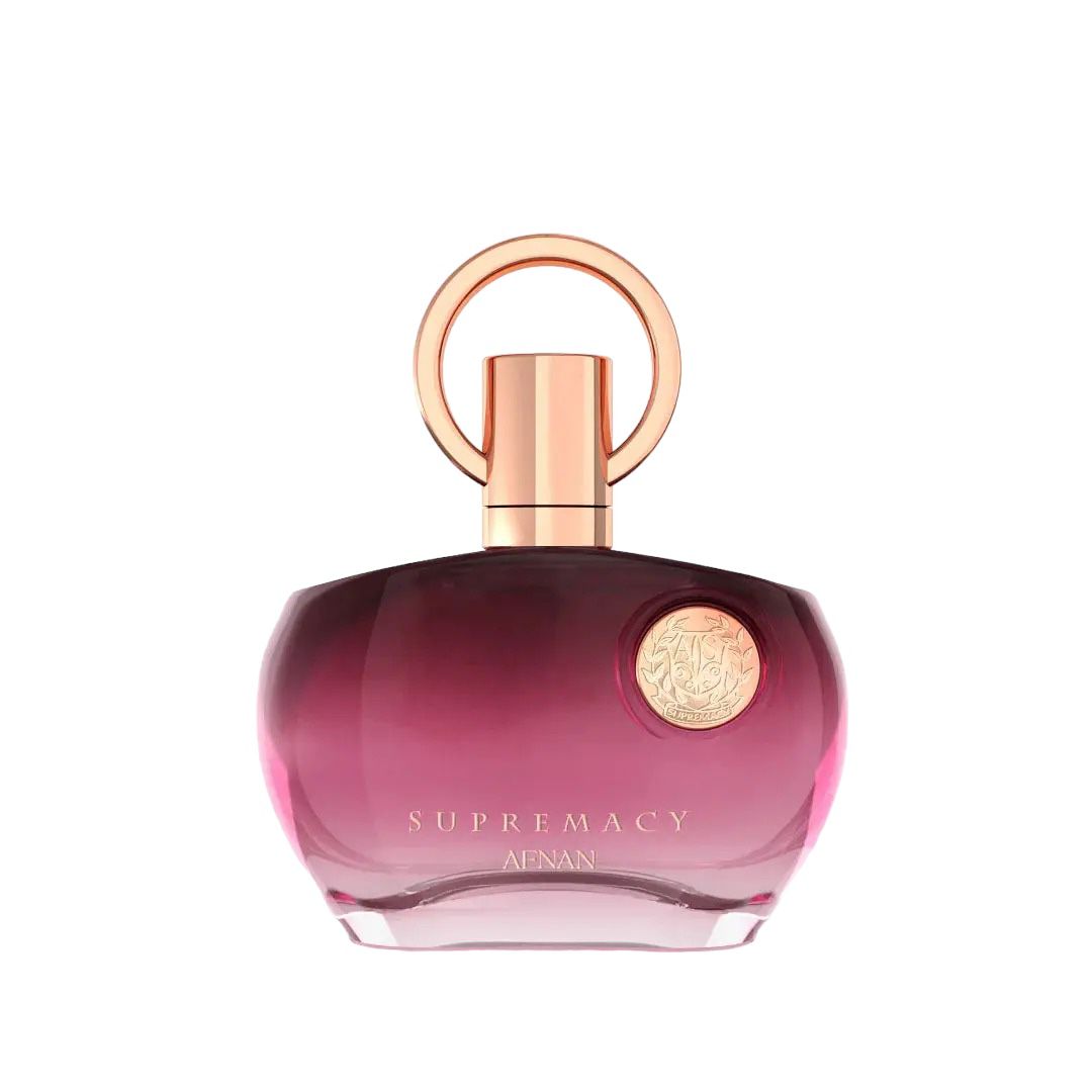 The image shows a perfume bottle with a distinctive design. It has a round, flat base with a gradient color that shifts from transparent to a deep pink hue towards the bottom. The cap is a metallic rose gold color, featuring a simple yet elegant circular design that loops around the nozzle. The front of the bottle has a metallic emblem with intricate detailing, adding a touch of luxury. The text on the bottle reads "SUPREMACY AENAN". 