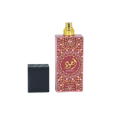 Image of a tall pink perfume bottle with a gold spray nozzle, placed upright next to its black textured cap which is lying on its side. The bottle has a decorative pattern in a lighter pink hue with a central circular label containing gold Arabic calligraphy and the word 'AJWAD'. Below the pattern, 'Lattafa' is written in small gold letters. The bottle is reflected on a glossy surface below, indicating a polished presentation.