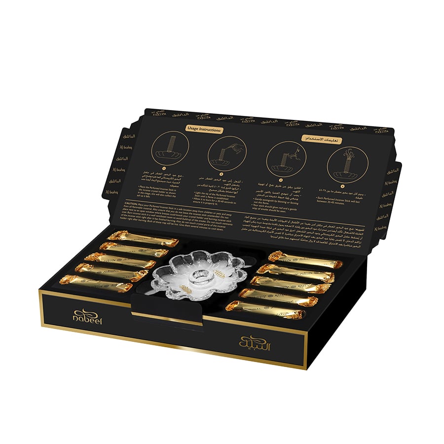 An open box of Al Bashiq Nabeel Perfumes, 50 grams, displayed with the lid propped up to reveal the contents and usage instructions. The box interior is black with gold accents and contains several gold-foil wrapped incense sticks, organized in neat rows, alongside a clear, flower-shaped incense burner in the center. The lid features detailed usage instructions illustrated with line drawings and written in both Arabic and English.