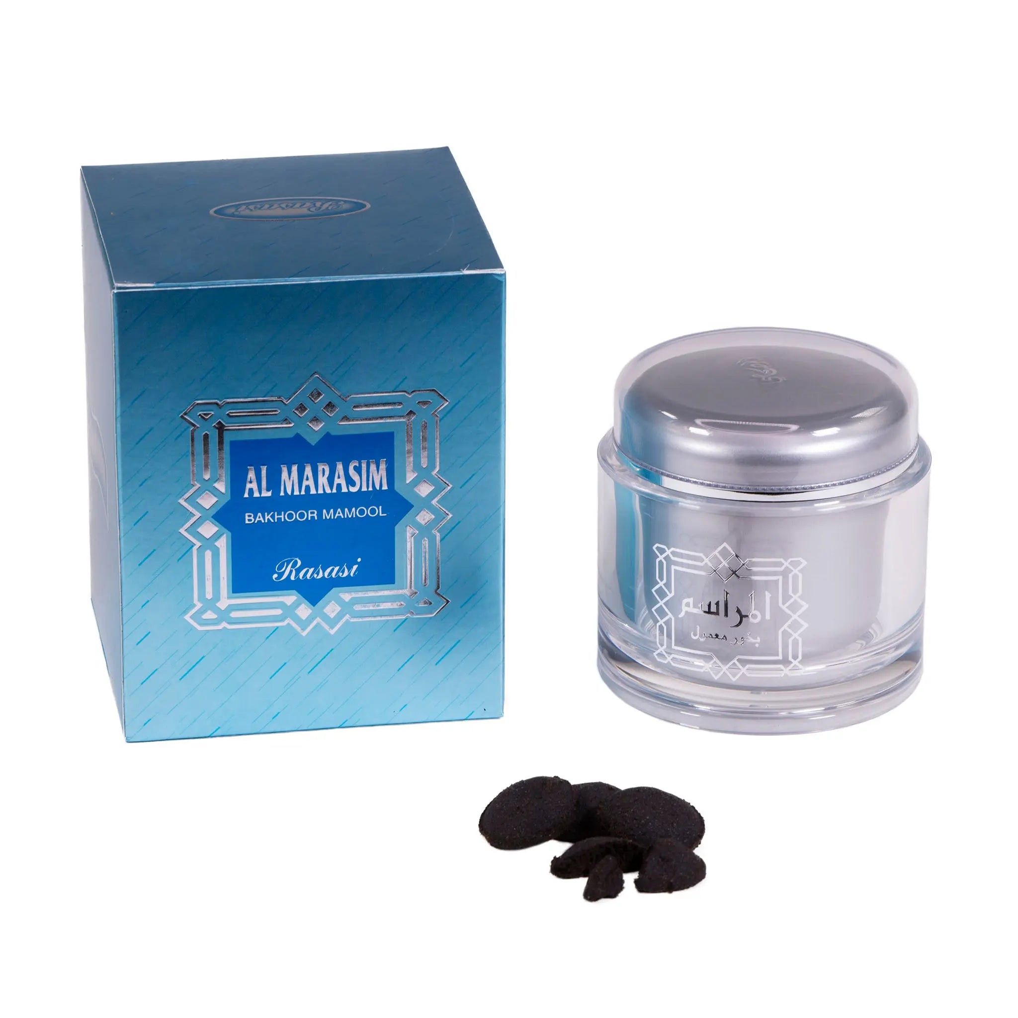 The image shows a product set that includes a jar of bakhoor incense, several pieces of the solid incense outside the jar, and the product's packaging box. The jar is made of clear glass with a silver lid and features Arabic and English writing that reads "Al Marasim Bakhoor Mamool" by "Rasasi." The packaging box is blue with a similar design and branding. The bakhoor pieces are dark brown and irregularly shaped.