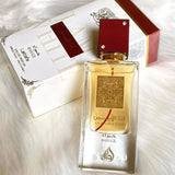 Image of a clear glass perfume bottle with a pale gold liquid, a red cap with two crystal embellishments, and a gold label featuring arabesque designs and Arabic script '??? ????' (Ana Abiyedh), with 'ROUGE' in red below. The bottle is placed at an angle on a white fluffy surface, with its white packaging box in the background. The box has the perfume's name and 'Eau de Parfum' printed on it, with a maroon stripe and golden floral motifs, suggesting a luxurious and elegant product.