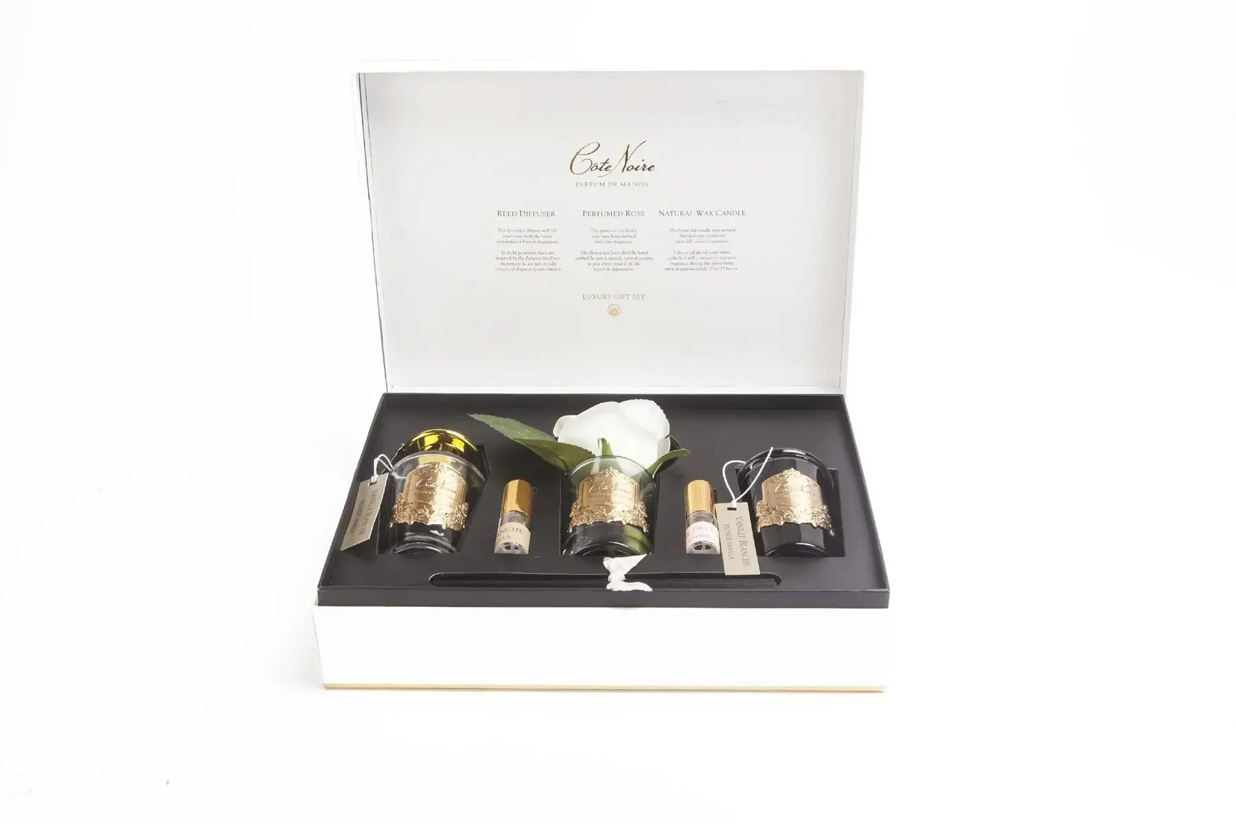 The image displays an open luxury gift set from Cote Noire. Inside the white box with gold edges, the set includes three glass jars with gold labels, each containing either a reed diffuser, a perfumed rose, or a natural wax candle. A single white rose with a stem is also prominently displayed in the center of the box. Each jar is tagged with a small, elegant label that describes the contents. The inner lid of the box contains descriptive text about each item in the set.