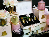 The image displays a luxurious and opulent Cote Noire gift set arrangement in a richly decorated setting. The main focus is a white box with gold trim, open to reveal a selection of fragrance products: two small perfume bottles, a natural wax candle, and a reed diffuser, all set against a black velvet background. Accompanying the box are various elegant elements, including a golden vase with white roses, a pink glass candle holder with a tassel.