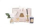 The image presents an elegantly arranged Cote Noire luxury gift set on a white background. The set includes a white gift box with a gold crest and is tied with a white bow. To the left, a single white rose in a clear glass vase with a gold label enhances the display. Beside the rose, two small bottles of perfume with gold caps and a large reed diffuser with a golden top and black reeds are showcased. On the far right, a dark blue glass candle with a gold label adds a striking contrast.
