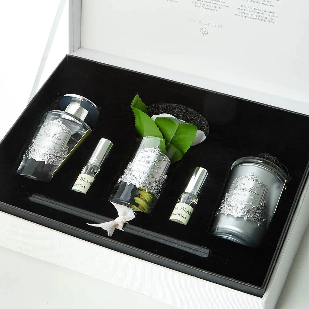 An open luxury gift set box from Côte Noire showcasing various items arranged in a black velvet-lined interior. The set includes a clear glass vase with an artificial white rose and green leaves, a reed diffuser with black reeds, two small glass bottles labeled 'Gardenia Blanc,' and a clear glass candle with a lid. Each item features an ornate silver label, and the box interior lid displays the 'Côte Noire Luxury Gift Set' branding in an elegant font.