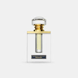 The image features a clear glass perfume bottle with a simple, elegant design. The bottle has straight lines with a rectangular body, allowing the light yellow-colored perfume inside to be visible. It is topped with a large, round cap that appears to be made of crystal-clear material, mounted on a gold band that adds a touch of luxury.