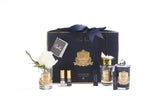 The image depicts a luxurious gift set from Côte Noire, elegantly presented on a white background. In the center, there is a dark blue box with a gold emblem and a navy ribbon, which holds various fragrance products. Arranged around the box are two glass votives with gold labels and a single artificial white rose in each, a perfume bottle with a gold sprayer, a smaller perfume vial, and a black diffuser stick, all bearing the distinctive Côte Noire branding.