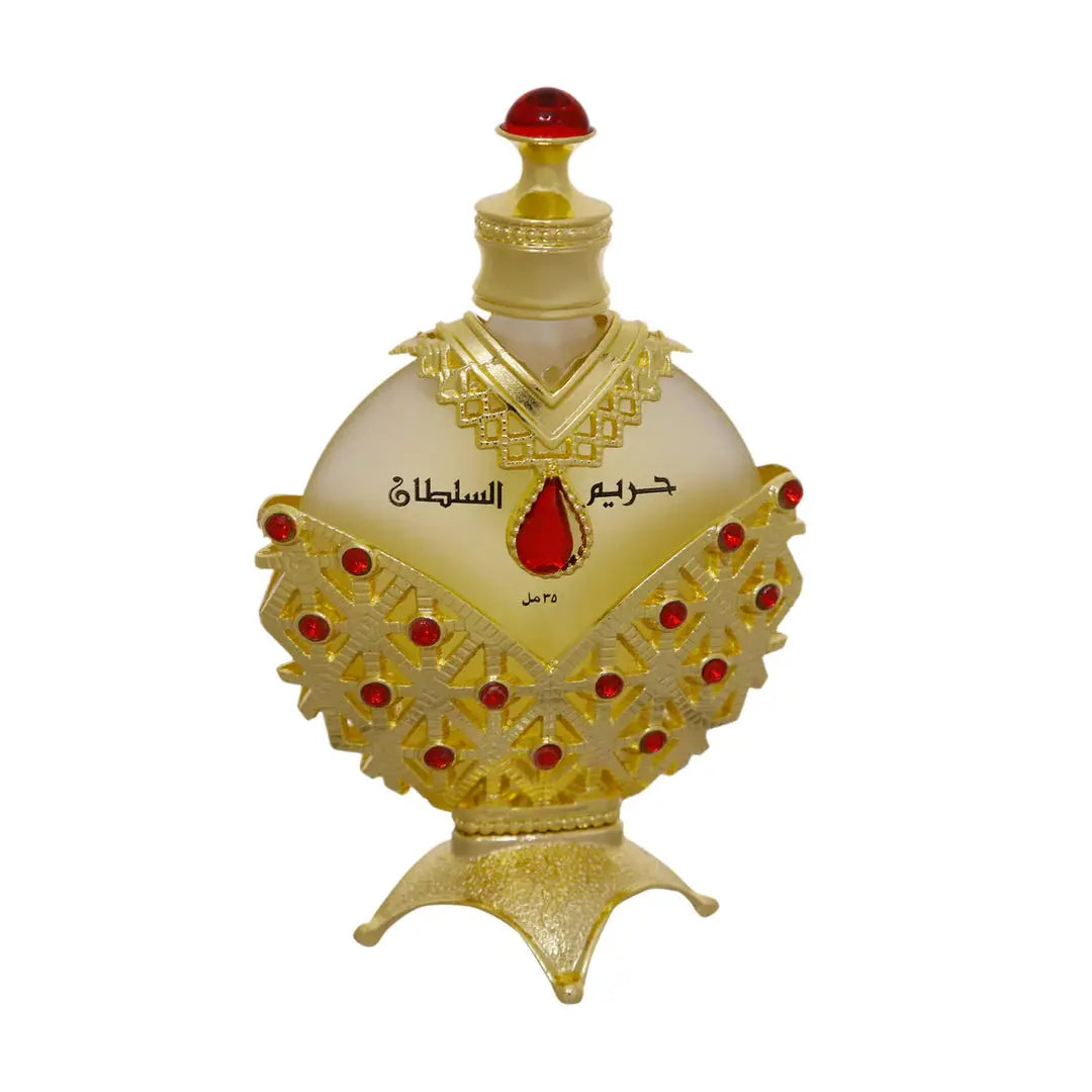 The image displays an opulent perfume bottle with a white background. The body of the bottle is a pale, translucent yellow, topped with a bright red cap resembling a gemstone. The bottle is adorned with a golden overlay featuring intricate cut-out patterns and red gemstone accents. Arabic script is prominently displayed in the center, with smaller text below.