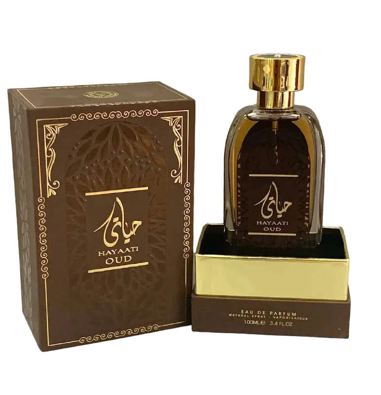n image of Hayaati Oud perfume by Ard Al Zaafaran, featuring the perfume bottle and packaging. The bottle is transparent with a dark amber liquid and a reflective gold cap. It sits on a gold-colored base with the text "EAU DE PARFUM NATURAL SPRAY 100ml e 3.4fl.oz" at the bottom. The brown box has an ornate design with golden accents and Arabic calligraphy, along with the name "HAYAATI OUD" in both Arabic and English.