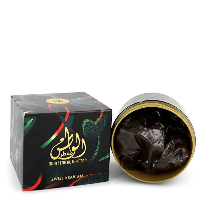 The image displays a product called "Muattar Al Watan" by "Swiss Arabian". It shows a cube-shaped black box with colorful abstract designs and both Arabic and English branding. Next to the box is an open container with a gold interior, revealing a black plastic bag that likely contains the fragrant substance. The product's packaging gives off a contemporary yet traditional vibe, with the bold calligraphy contrasting with the modern design elements on the box.