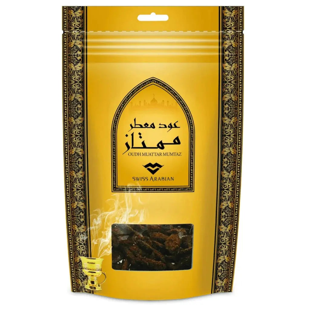 The image shows a package of "Oudh Muattar Mumtaz" by Swiss Arabian. The packaging is bright yellow with intricate black and gold floral border designs. Centered on the package is an arch-shaped window displaying Arabic calligraphy and the silhouette of a mosque in the background, giving it a distinctively Middle Eastern look. Below the arch, there is a clear plastic window through which dark brown oud wood chips are visible, adding an authentic touch. 