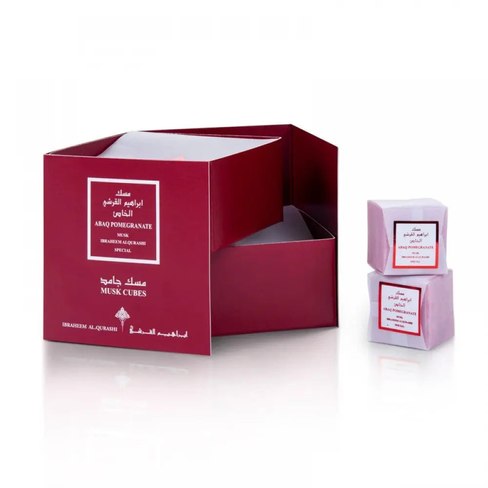 The image shows a product display consisting of a deep red open box labeled in both English and Arabic. Inside the box are small pink cubes, which are also individually labeled. The main label on the box reads "ABAQ POMEGRANATE" and "MUSK CUBES", indicating the scent and product type, and includes "IBRAHEEM AL QURASHI SPECIAL". Two cubes are placed in front of the box, one stacked on top of the other, to showcase the product's packaging and presentation.