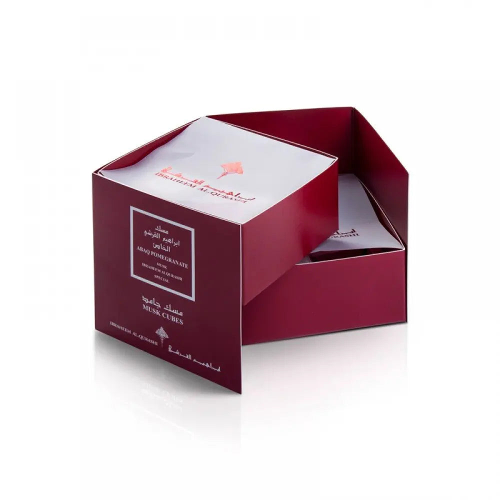 A photograph of a partially open dark red box with white interior, labeled "ABAQ POMEGRANATE" and "MUSK CUBES" in white lettering, also including Arabic script and the brand "IBRAHEEM AL QURASHI". The product name suggests the box contains scented musk cubes with a pomegranate fragrance. The box lid, which is visible inside the box, has an illustration of a pomegranate.