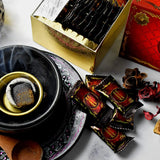 The image is a stylized product display for "Bakhoor Nasaem" by Nabeel Perfumes. It shows a traditional black bakhoor burner with a lit piece of bakhoor inside, from which smoke is rising, placed on a decorative plate. In front of the burner are several small, sealed packets with a maroon and gold design, labeled with the "Nasaem" brand name, which contain bakhoor. To the right, a golden box with a reflective surface, partially visible, seems to be the packaging for the bakhoor packets.