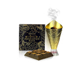 The image features a product presentation of 'Nabeel Bakhoor Black', which seems to be a type of scented bakhoor or incense. In the foreground, there are square-shaped bakhoor pieces with a pyramid-like emboss on each. To the right, there is a decorative gold-colored incense burner with smoke rising from the top, indicating the bakhoor is being used. Behind these items is the product packaging, a box with a black background and gold leaf designs, along with the text 'Bakhoor Nabeel Black'.