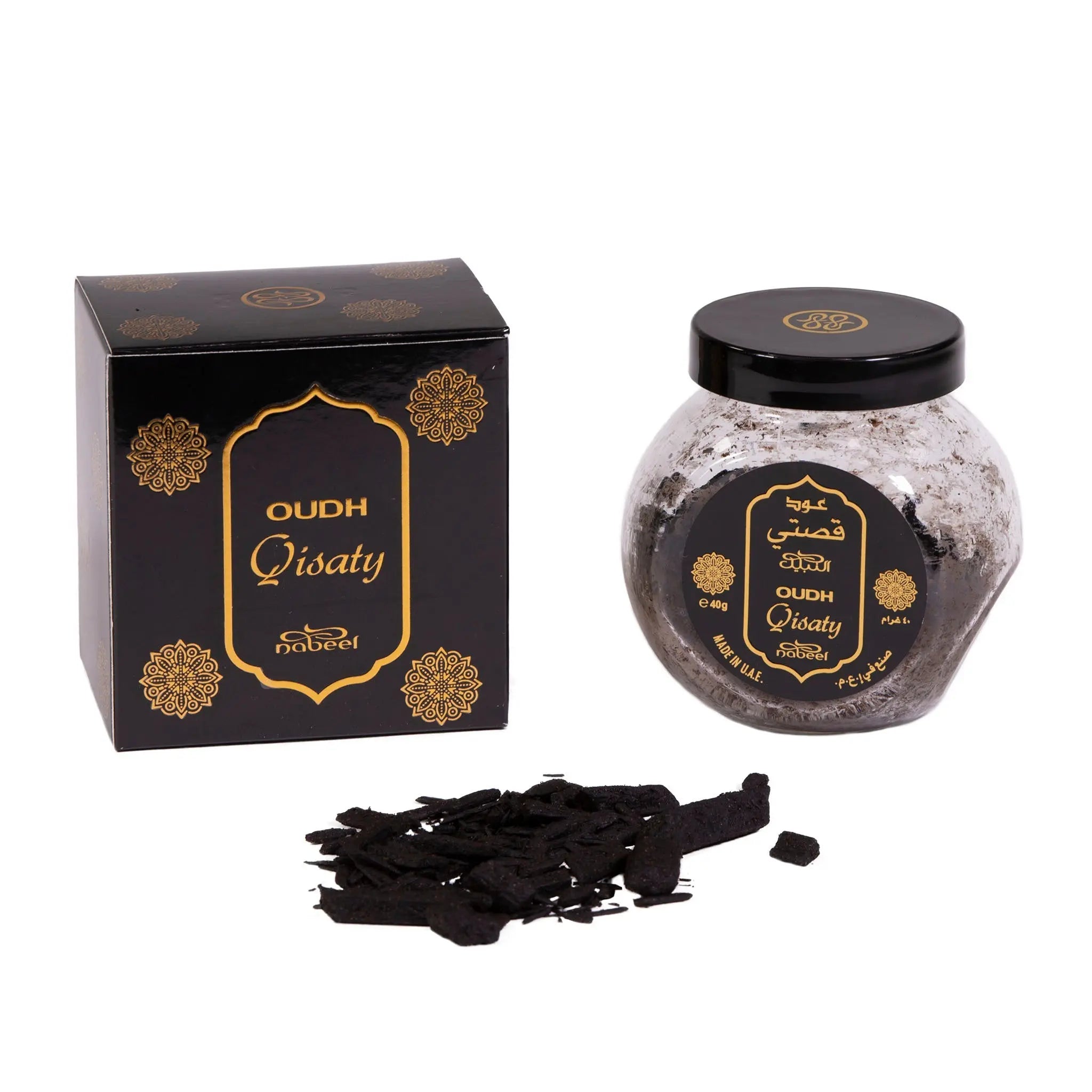 The image depicts "OUDH Qistaty" by Nabeel, presented with a black box and a clear glass jar, both adorned with gold floral accents and branding. The product name is displayed in both Arabic and English on a gold label. In front of the jar lies a scattered pile of dark, resinous oudh chips, which are used for their fragrant properties. The black lid of the jar matches the box, and both feature the Nabeel logo.