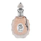 The image shows a decorative perfume bottle:  The bottle has an ornate, multi-faceted glass design with a textured surface and a pale pink tint, suggesting a delicate fragrance within. It features a clear glass stopper with an intricate cut pattern on top. A metallic silver chain with a charm and a small black stone is attached to the neck of the bottle. The front of the bottle displays a white oval label with Arabic calligraphy and the name "ROUAT AL MUSK" in Roman characters below.