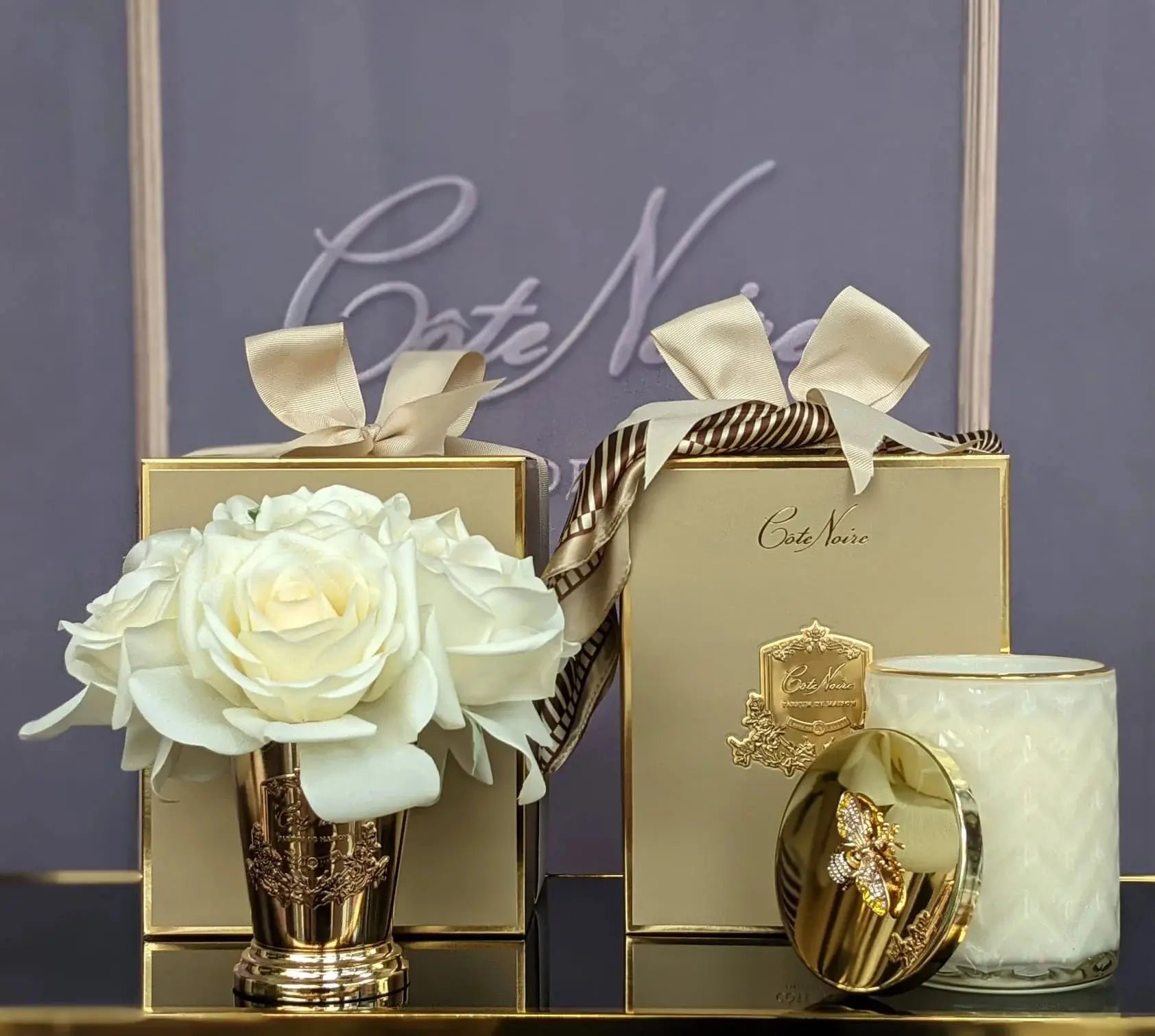 A Cote Noire luxury gift set featuring a champagne gold vase filled with white artificial roses, and two elegant gold gift boxes adorned with cream-colored ribbons. One box contains a white textured candle with a gold lid decorated with a jeweled bee. The items are displayed against a sophisticated background with the Cote Noire logo, creating a luxurious and refined presentation.