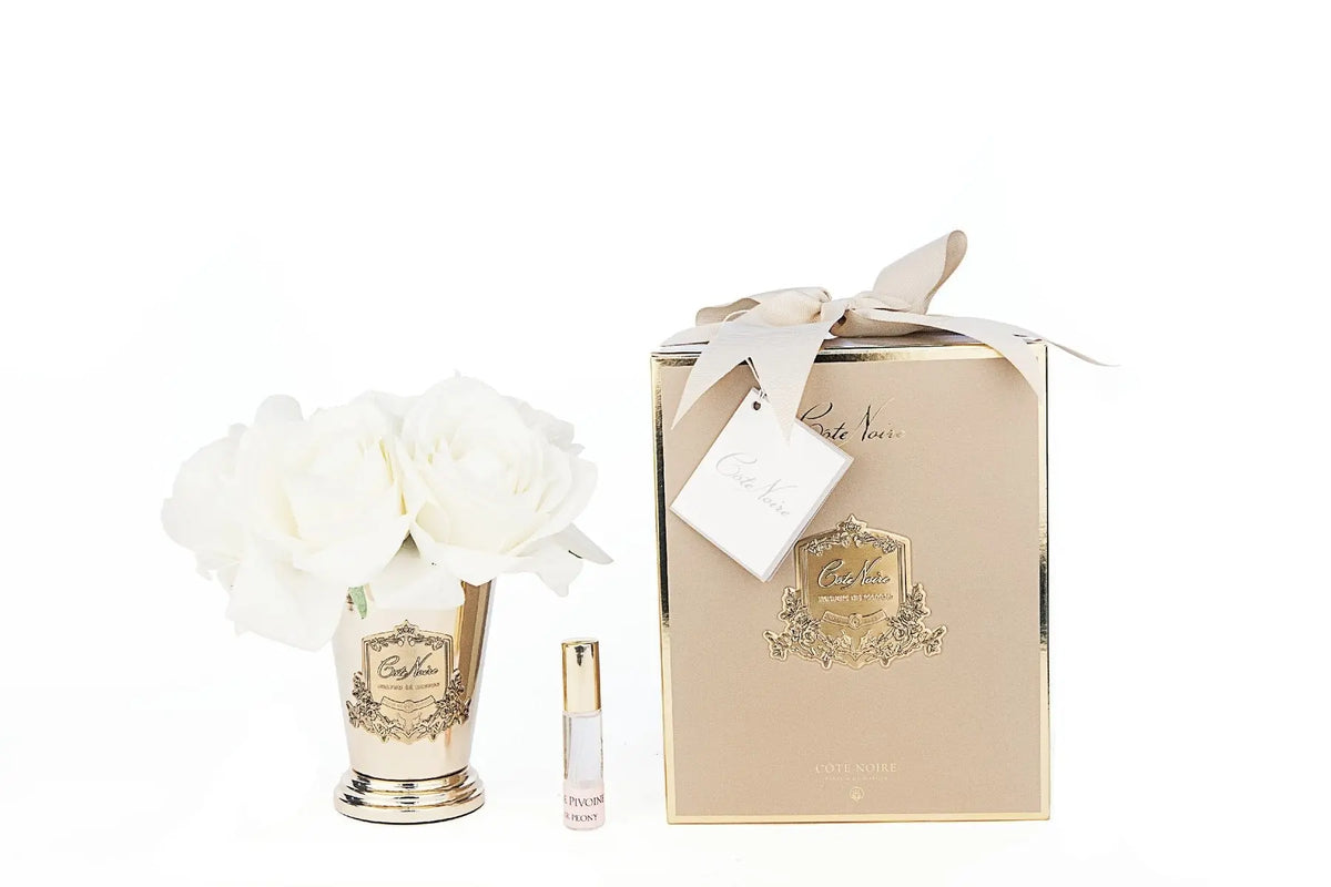 A Cote Noire gift set featuring a champagne gold vase filled with seven white artificial roses, alongside a small vial of perfume labeled 'Pétale de Rose' with a gold cap. The items are displayed next to a matching gold gift box adorned with a gold emblem and a cream-colored ribbon. The box includes a tag with the Cote Noire logo, creating a luxurious and elegant presentation.