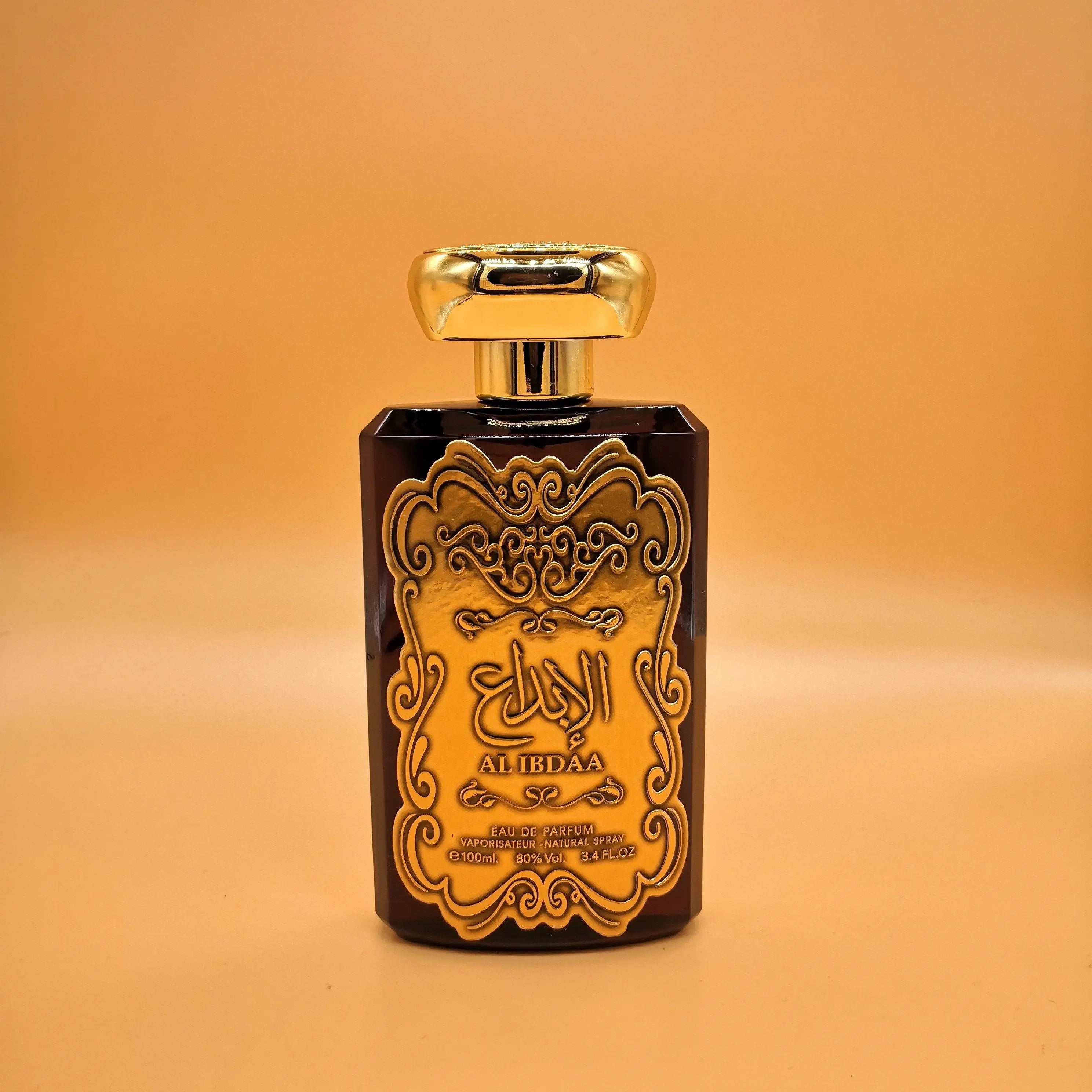 The image shows a perfume bottle with a dark, translucent body and a shiny gold label. The label features intricate, ornate designs and Arabic calligraphy that reads "AL IBDAA," which is likely the name of the fragrance. Below the calligraphy, in smaller English text, it says "EAU DE PARFUM NATURAL SPRAY 100ml 3.4FL.OZ," indicating the contents and volume. The cap of the bottle is gold and has a simple, elegant shape. 