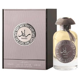 The image shows a perfume bottle next to its packaging box. The bottle is transparent with a golden liquid inside and features a brown cap and a label with Arabic calligraphy and English text "RA'ED Lattafa". A brown tassel is attached to the bottle's neck. The square box is colored in shades of grey and black with a circular design in the center that matches the bottle's label, displaying the same Arabic calligraphy and English text.