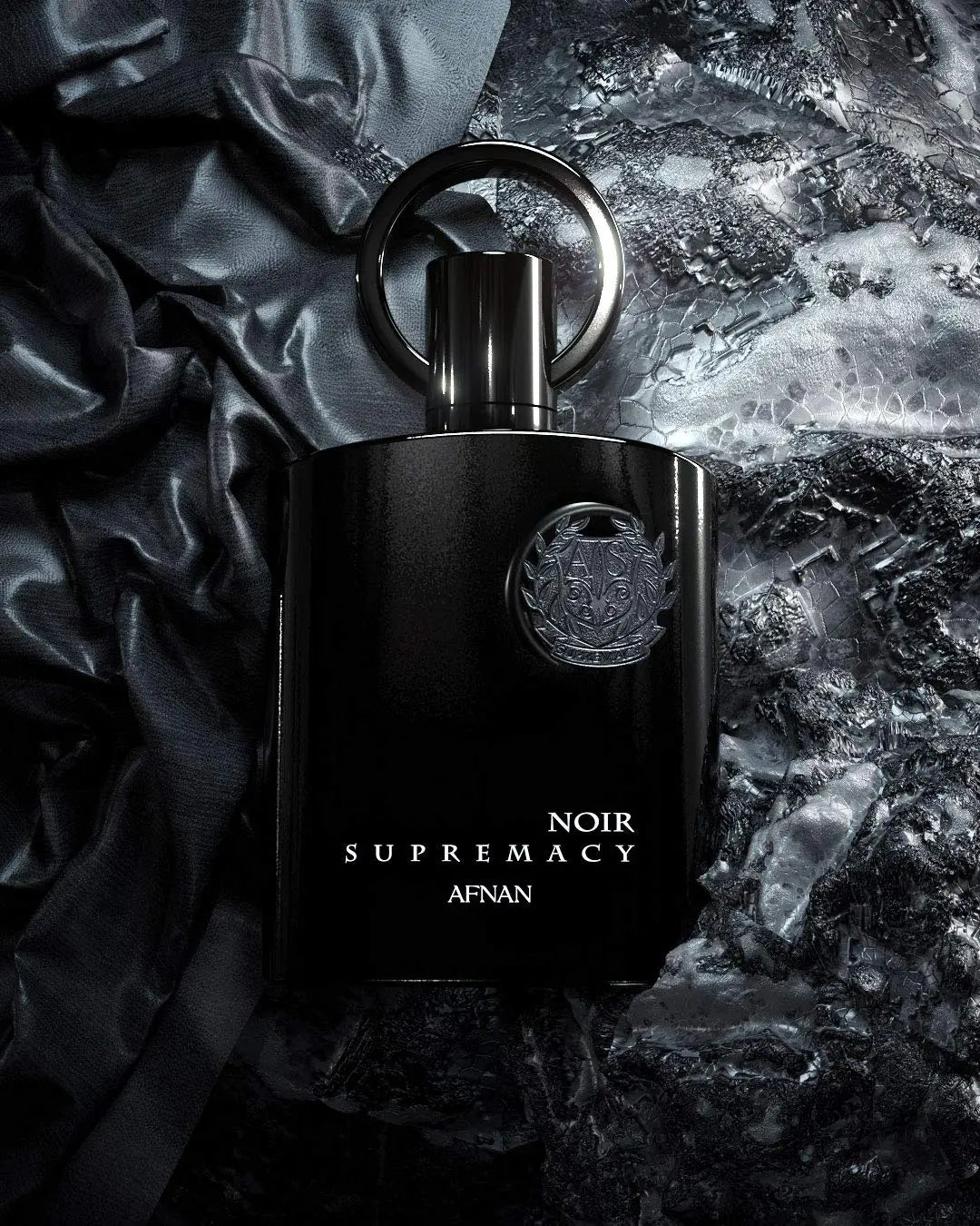 The image depicts a black perfume bottle labeled "NOIR SUPREMACY AFNAN" resting on a textured surface that appears to be a mix of crumpled satin fabric and reflective metallic foil. The perfume bottle has a unique design with a ring-shaped cap at the top and features a prominent metallic emblem. The composition of the photo creates a luxurious and dramatic effect, with the dark tones and textures emphasizing the sleekness and elegance of the perfume bottle.