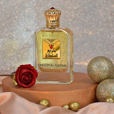 This image presents a perfume bottle named HAREEM AL SULTAN by Khadlaj. The perfume bottle is clear glass with a golden-yellow fragrance liquid inside. It features a decorative gold cap and a gold label with the name in both Arabic calligraphy and English, adorned with a graphic of a red gemstone. The setting includes a red rose and golden glittery ornaments on a wooden surface, with a sparkly background that adds a festive or luxurious feel. 