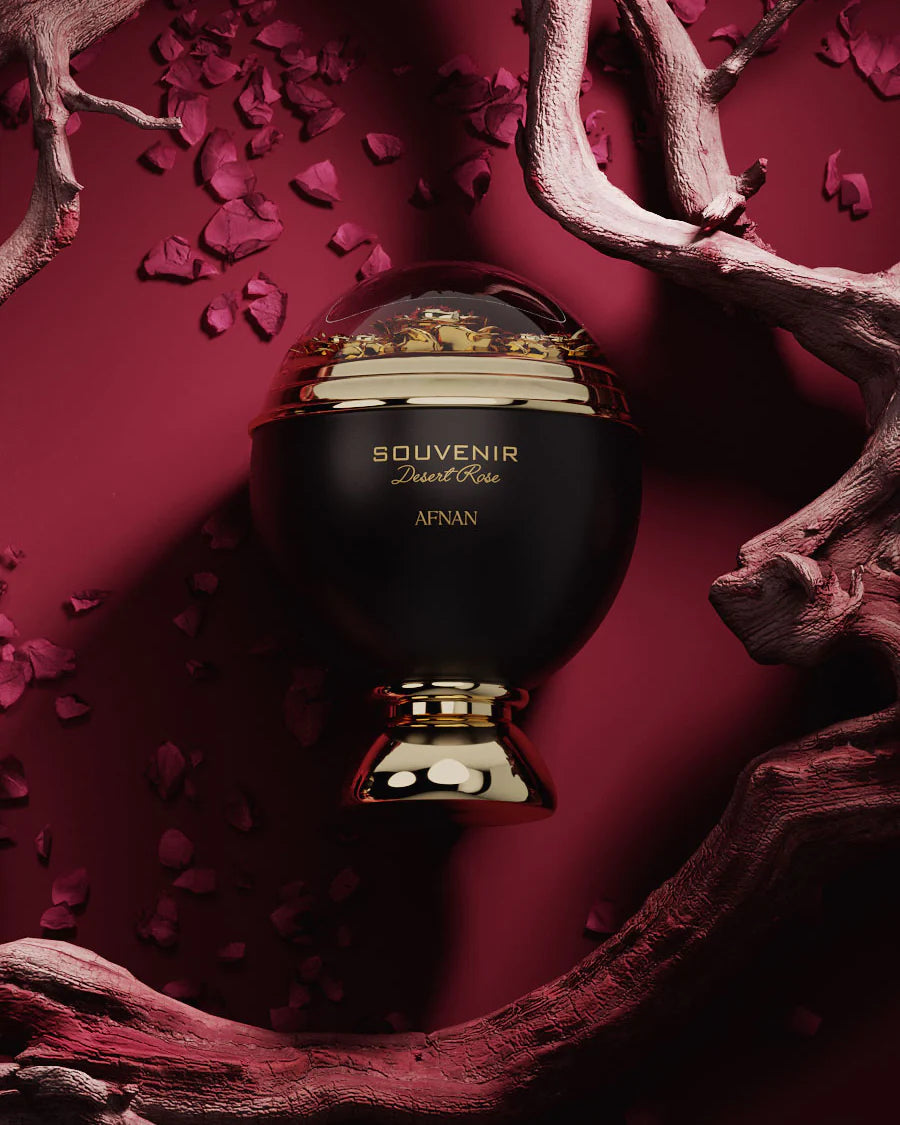 The image features a perfume bottle presented in a dramatic setting. The bottle, titled "SOUVENIR Desert Rose AFNAN," has a matte black spherical body and a transparent dome on top, within which gold flakes are visible, creating a luxurious visual effect. A golden band encircles the bottle, matching the elegant golden pedestal on which it stands.