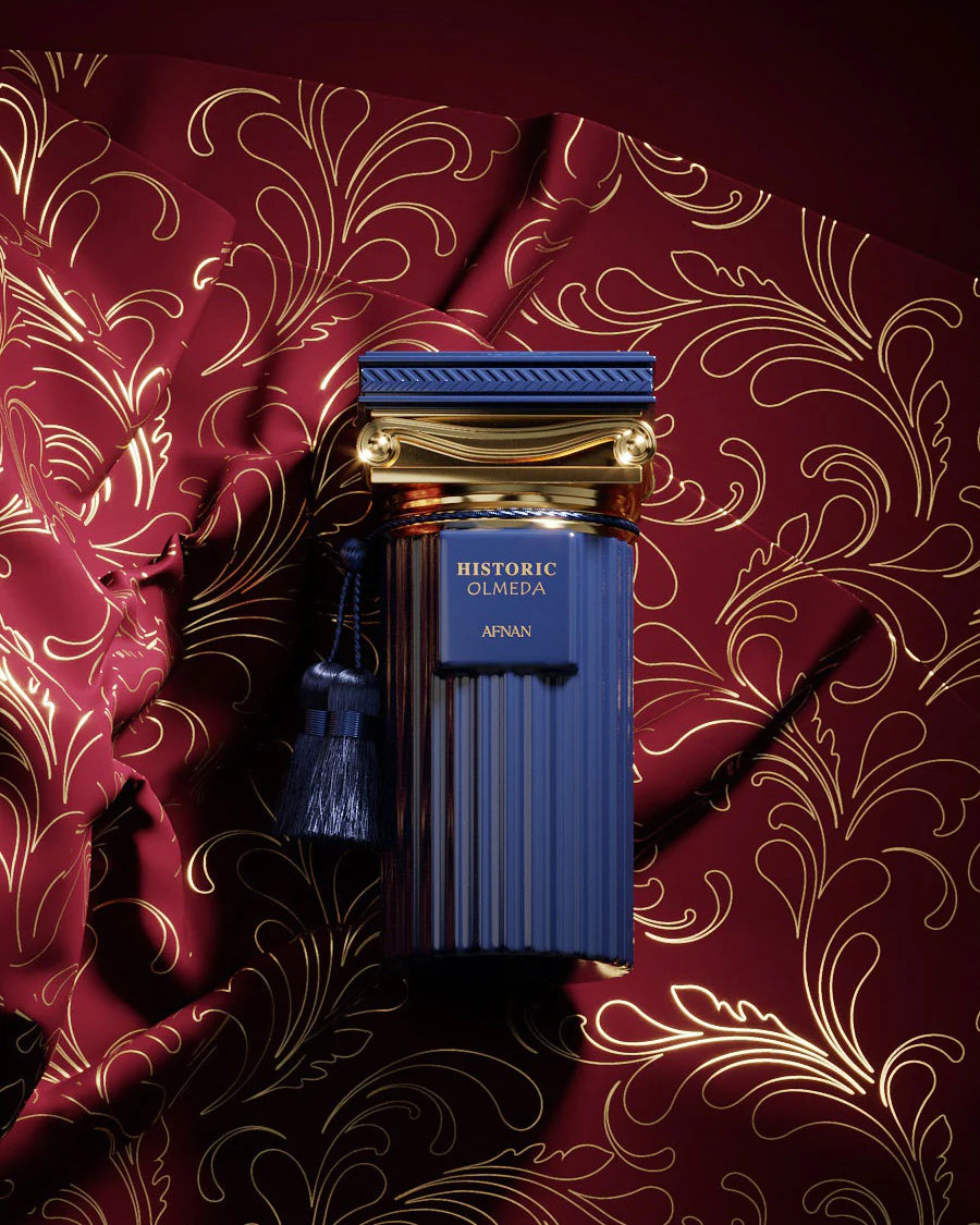 The image features a luxurious fragrance bottle placed against a richly patterned background. The bottle has a deep blue color with vertical ridges and is adorned with a gold cap that has a classical design, complete with a blue tassel. The gold label on the bottle reads "HISTORIC OLMEDA AFNAN." The backdrop is a dark red fabric with a glossy finish, embellished with a gold floral pattern that exudes opulence and elegance.