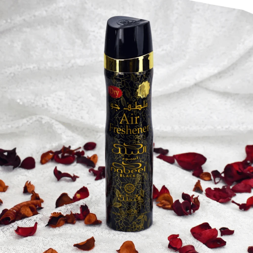 The image features a cylindrical can of 'Nabeel Black' air freshener. The can has a black background with gold and white Arabic calligraphy and English text, including the words "Dry Air Freshener." There's a decorative golden band around the top of the can. Surrounding the can are scattered dried red rose petals on a textured white fabric surface, adding to the aesthetic appeal and suggesting a floral scent.