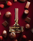 The image presents a perfume bottle named "Mirsaal with love" by Afnan Perfumes, set against a luxurious backdrop of chocolate treats and strawberries. The bottle has a clear, geometric design with a gold neck and a textured wooden cap, conveying a sense of warmth and elegance. The background is a deep, rich red, complementing the golden accents of the bottle and the assortment of chocolates and strawberries scattered around it, hinting at the perfume's possibly sweet and indulgent fragrance notes. 