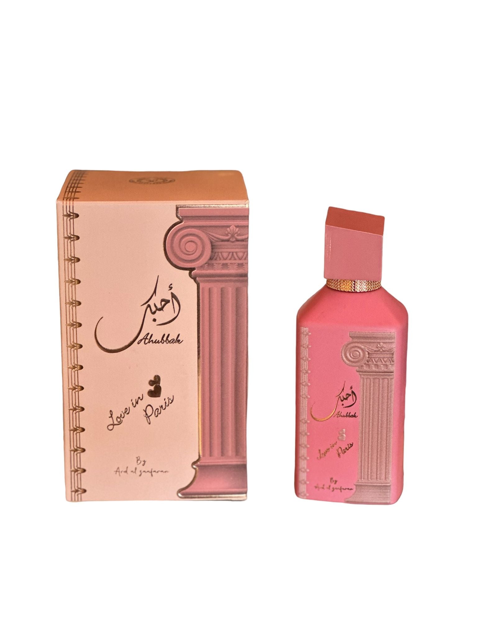 The image shows a set of two products with a cohesive design theme. On the left is a notebook or journal, and on the right appears to be a bottle, possibly of perfume. Both items are primarily in a pastel pink hue. The notebook features a graphic of a classical column along the binding edge, giving it an elegant look, with script writing that suggests the theme "Love in Paris." There is also a signature or designer's mark in a foreign script, possibly Arabic, near the top.
