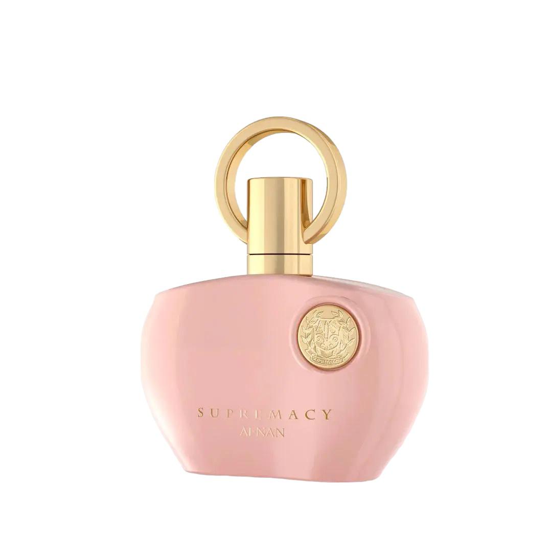 The image displays a perfume bottle from Afnan's "SUPREMACY" line. The bottle is made of smooth, matte pink glass and has a rounded, organic shape with a flat base, allowing it to stand upright. The cap is a glossy golden hue, designed with a loop that creates a visual focus and adds an element of luxury. On the front of the bottle is a golden emblem, adding a touch of elegance to the overall design. Below the emblem, in a simple and sophisticated font, reads "SUPREMACY AFNAN." 