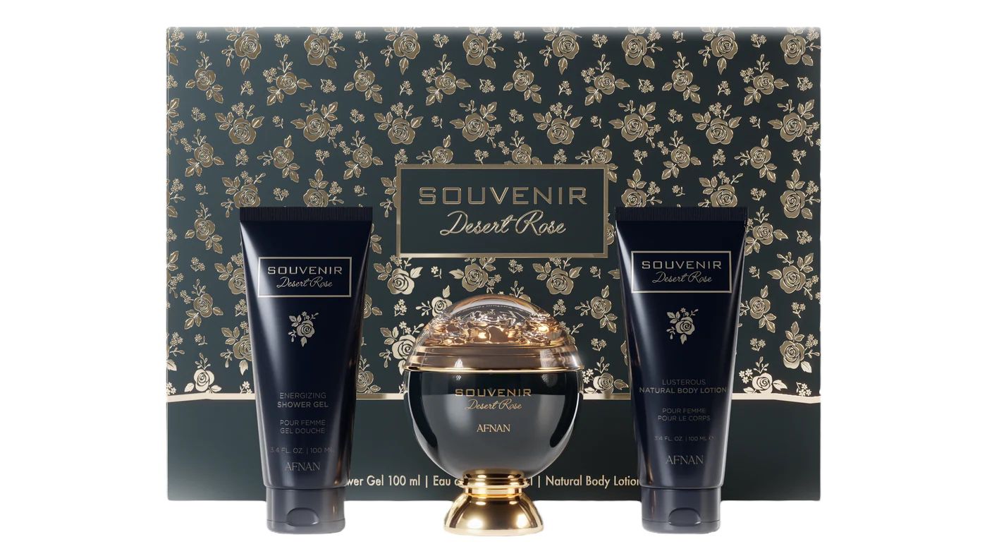 The image features a luxurious bath and body set from the "SOUVENIR Desert Rose" collection by Afnan. The set includes a spherical golden perfume bottle with a clear dome top, flanked by two navy blue tubes—one labeled as "ENERGIZING SHOWER GEL" and the other as "LUSTEROUS NATURAL BODY LOTION". Each product has gold lettering and rose accents. The background and base of the set feature an elegant pattern of gold roses on a dark, forest green backdrop.