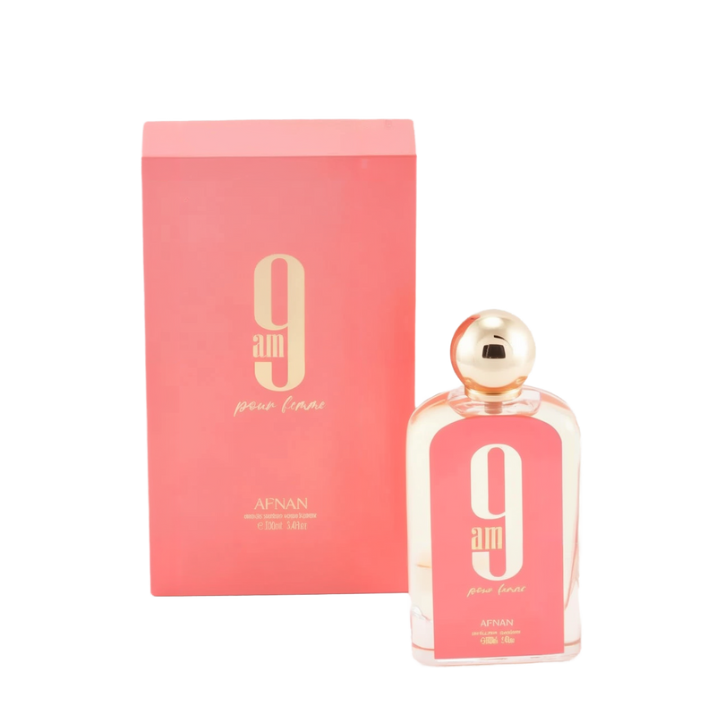 The image displays a rectangular glass perfume bottle and its corresponding box. The bottle is clear with a pink hue and has a white oval label with a large "9" and "am" in black font, with "pour femme" in smaller script below it. The cap is a polished gold sphere. At the bottom of the bottle, "AFNAN" is printed, along with "100ml | 3.4fl.oz". The box is a matching pink color with the same "9 am pour femme" branding and additional text "eau de parfum vaporisateur". 