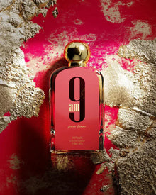 The image features a rectangular glass perfume bottle with a pink to red gradient background. The bottle has a white oval label with a large black "9" and below it "am" in a matching color, with "pour femme" in smaller script beneath that.  "AFNAN" is printed at the bottom of the bottle along with the volume "100ml | 3.4fl.oz". The backdrop is striking, with an abstract texture of gold leaf overlaid on a vibrant red paint, giving the impression of luxury and boldness.