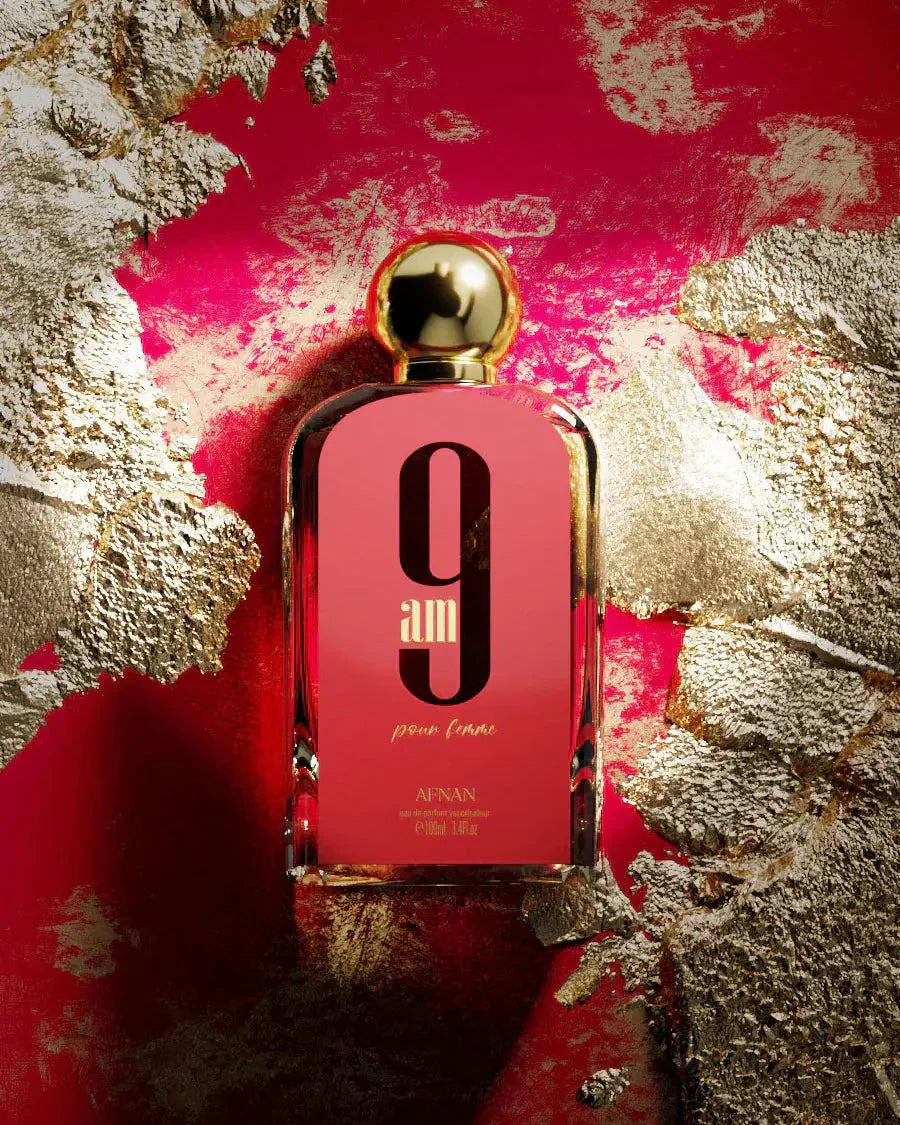 The image features a rectangular glass perfume bottle with a pink to red gradient background. The bottle has a white oval label with a large black "9" and below it "am" in a matching color, with "pour femme" in smaller script beneath that.  "AFNAN" is printed at the bottom of the bottle along with the volume "100ml | 3.4fl.oz". The backdrop is striking, with an abstract texture of gold leaf overlaid on a vibrant red paint, giving the impression of luxury and boldness.