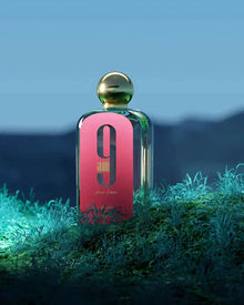 The image shows a rectangular glass perfume bottle with a pink front label and a gold spherical cap, placed on a surface with greenery. The label features a large black "9" and "am" in black font, with "pour femme" in smaller script below it. The text "AFNAN" is visible at the bottom of the label.  The bottle is set against a moody background with a blue gradient, evoking a night-time scene, and is illuminated from above, creating a soft reflection on the cap.