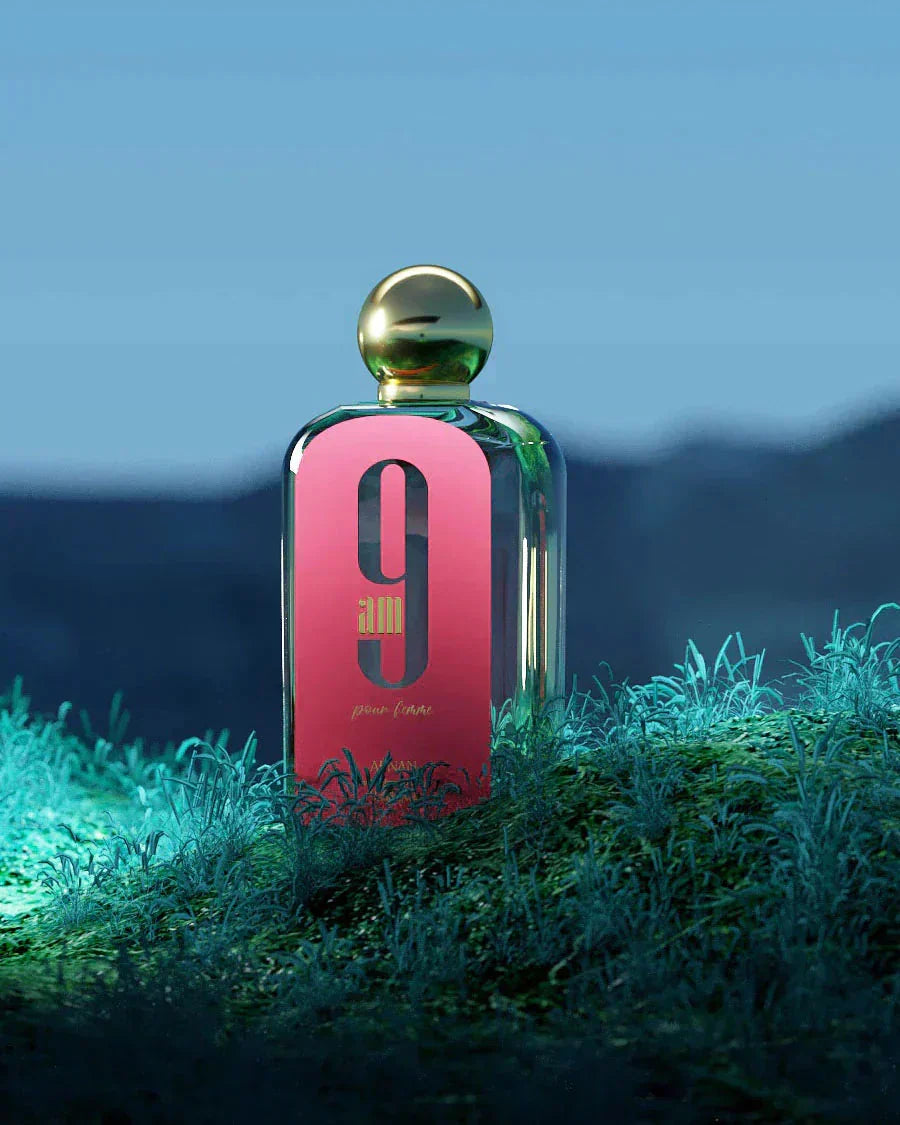 The image shows a rectangular glass perfume bottle with a pink front label and a gold spherical cap, placed on a surface with greenery. The label features a large black "9" and "am" in black font, with "pour femme" in smaller script below it. The text "AFNAN" is visible at the bottom of the label.  The bottle is set against a moody background with a blue gradient, evoking a night-time scene, and is illuminated from above, creating a soft reflection on the cap.