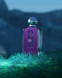 The image depicts a perfume bottle placed on a grassy surface with a blurred natural background suggestive of a twilight setting. The bottle has a sleek, rectangular shape with a metallic spherical cap. It features a vibrant purple label with the white text "9 pm pour femme" in a bold font, and the brand "AFNAN" is visible at the bottom of the label. The overall atmosphere of the image is moody and atmospheric, likely to emphasize the scent's evening wear suitability.