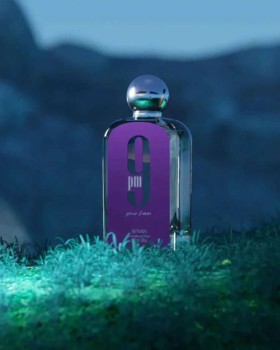 The image depicts a perfume bottle placed on a grassy surface with a blurred natural background suggestive of a twilight setting. The bottle has a sleek, rectangular shape with a metallic spherical cap. It features a vibrant purple label with the white text "9 pm pour femme" in a bold font, and the brand "AFNAN" is visible at the bottom of the label. The overall atmosphere of the image is moody and atmospheric, likely to emphasize the scent's evening wear suitability.
