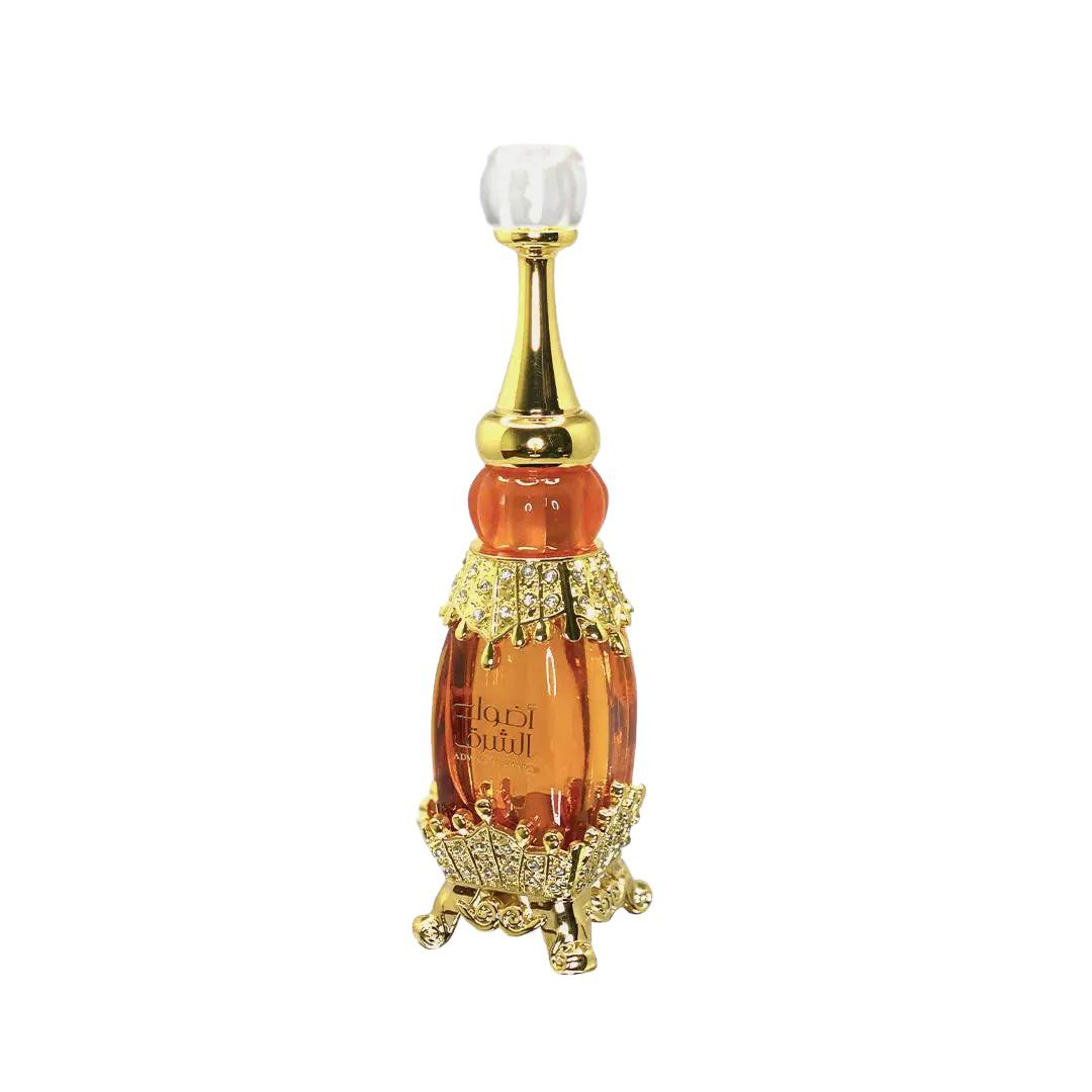 The image shows a luxurious perfume bottle from the brand "Adwaa Al Sharq." The bottle has a tall, elegant design with a combination of amber and gold hues. It features a crystal-like cap, a golden neck, and intricate gold embellishments encrusted with small rhinestones. The base of the bottle is also adorned with ornate gold details, giving it an opulent and sophisticated appearance.