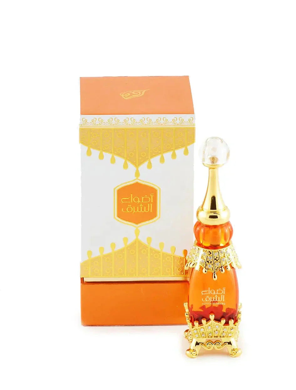 The image displays a luxurious perfume bottle from the brand "Adwaa Al Sharq" alongside its packaging. The bottle has an elegant design with amber and gold colors, a crystal-like cap, and intricate gold embellishments encrusted with small rhinestones. The packaging is predominantly orange and white, with a decorative gold pattern and the brand name "Adwaa Al Sharq" prominently displayed on the front. 