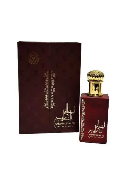 The image showcases a premium perfume bottle nestled within a bed of golden sand and illuminated by warm, soft lighting. The perfume bottle has an elegant design with a deep red, glossy finish. Its body is adorned with intricate golden patterns and calligraphy that suggest a Middle Eastern or Arabian influence. The label on the bottle reads "Ahlam Al Khaleej Eau de Parfum." The golden cap of the bottle is richly textured and adds to the luxurious appearance. 
