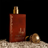 The image displays a luxurious perfume bottle on a bed of sparkling crystals. The bottle has a matte, earthy red finish with intricate golden scrollwork patterns along one side. Arabic calligraphy and the words "Ahlam Al Khaleej Eau de Parfum" are elegantly inscribed on the front in gold, suggesting the perfume's name and origin. Beside the bottle lies its cap, which has a gold finish with a ribbed texture, resembling a traditional incense burner or a decorative element with cultural significance. 