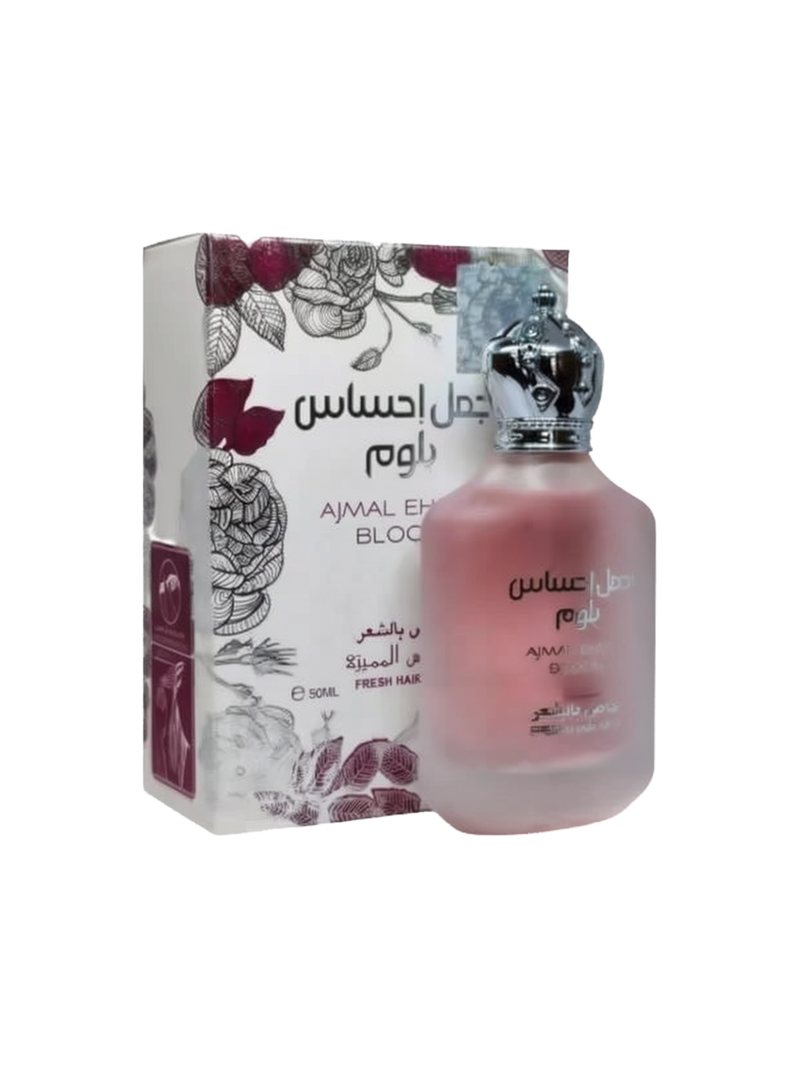 The image depicts a frosted glass perfume bottle with a pink gradient and a metallic cap, next to its packaging box. The box has a white background with a detailed floral design in shades of red and grey, and text that includes "AJMAL EHSAS BLOOM" along with Arabic script. The bottle features the same name in both English and Arabic, and additional text likely indicating the fragrance type and volume, "50ML FRESH HAIR MIST." 