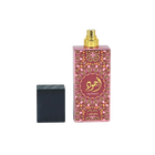 Image of a tall pink perfume bottle with a gold spray nozzle, placed upright next to its black textured cap which is lying on its side. The bottle has a decorative pattern in a lighter pink hue with a central circular label containing gold Arabic calligraphy and the word 'AJWAD'. Below the pattern, 'Lattafa' is written in small gold letters. The bottle is reflected on a glossy surface below, indicating a polished presentation.