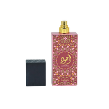 Image of a tall pink perfume bottle with a gold spray nozzle, placed upright next to its black textured cap which is lying on its side. The bottle has a decorative pattern in a lighter pink hue with a central circular label containing gold Arabic calligraphy and the word 'AJWAD'. Below the pattern, 'Lattafa' is written in small gold letters. The bottle is reflected on a glossy surface below, indicating a polished presentation.