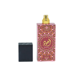 Image of a tall pink perfume bottle with a gold spray nozzle, placed upright next to its black textured cap which is lying on its side. The bottle has a decorative pattern in a lighter pink hue with a central circular label containing gold Arabic calligraphy and the word 'AJWAD'. Below the pattern, 'Lattafa' is written in small gold letters. The bottle is reflected on a glossy surface below, indicating a polished presentation.